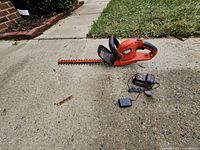 Hedge trimmer, battery and charger laid out on pavement
