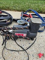 Portable compressor with gauge and braided hose, jumper cables in background