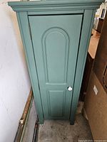 Front view of tall narrow sage/teal cabinet with single recessed panel door and white knob