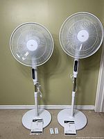 Two white HOMCOM pedestal fans side by side, full view with bases, remotes and manuals visible