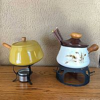 Two enamel fondue pots on stands; yellow pot left, cream wheat design pot right, five skewers visible