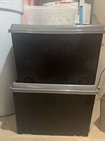 Two stacked black storage totes with grey lids, clear bin partially visible