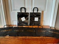 Front view showing five unwrapped bags laid flat spelling TOUCH, TASTE, SMELL, SIGHT, SOUND; additional packaged bags behind with ruler for scale