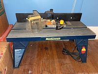 Overall view of router table showing aluminum slotted top, blue legs, fence, guard and power cord