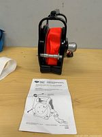 Side view of reel showing red strap inside black housing, axle visible, instruction sheet on table