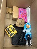 Top view of open shipping box showing various packaged items including Exvion box, Dhool boxes, rod holder, Master Plumber fitting, white wrapped item, black case and cylindrical rod
