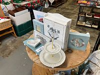 Overhead view of entire blue and white decor lot on round wooden table showing cabinet, risers, tiered tray, dish, plaque, frame and print