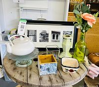 All items arranged on tabletop: teapot on cake stand, framed photo art, candlestick, glass bottle with flower, glass vase, ceramic dish, wooden floral box