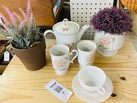 All items arranged on wooden surface showing teapot, two mugs, teacup with saucer, rose-pattern planter with faux foliage, grey pot with faux foliage