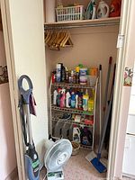 Overview of closet showing shelf filled with cleaning products, hangers, fan and handled tools