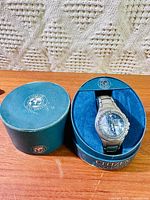 Watch seated in open teal Citizen Eco-Drive cylindrical box with lid beside it