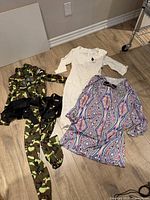 Three costumes laid out: white dress, patterned disco dress with scarf, camo jumpsuit with harness