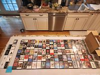 Overhead view of entire lot spread on countertop showing quantity and variety of cassette tapes plus storage cases box at right