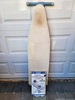 Full view of folding ironing board standing upright against garage door, Ironees cover package placed at base