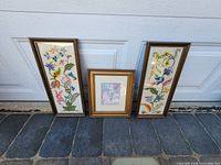 Three framed pieces standing: two tall floral needlepoints and central cat print