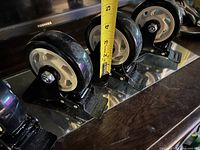 Four caster wheels on reflective surface showing size reference tape at ~3 in.