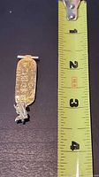 Front view of gold cartouche pendant beside tape measure showing length