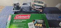 Two Coleman stoves (one assembled in case, one case closed), boxed stove, and griddle box visible