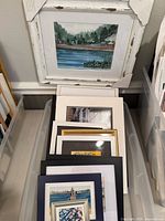 Stack of framed and matted artworks in storage bin showing variety of pieces and frames