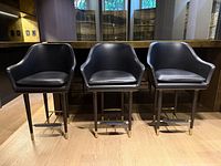 Front view of three black leather bar stools with oak legs and brass accents