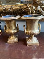 Front view of both gold ceramic urn planters on plinth bases