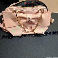 Front view showing pink faux-leather handbag with large bow, polka dots, gold chain, ruler indicating width