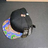 Side view of black cap with blue paisley brim, tags visible