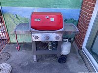Full view of Weber red-lid 3-burner gas grill with side shelves and propane tank shown for scale