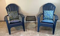 Two blue plastic Adirondack chairs, metal mesh table between, pillows displayed
