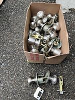 Open box on pavement containing numerous metal door knob assemblies with separate latch and strike hardware