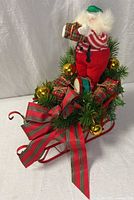 Full view of Santa figure on red metal sleigh with plaid gifts, greenery, gold ornaments and ribbon