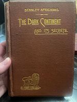 Front cover of ‘The Dark Continent and Its Secrets’ showing title, author attribution, gilt lion emblem