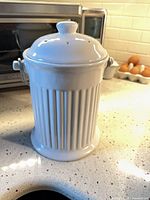 White ribbed ceramic compost bin with vented lid and side handles on kitchen counter