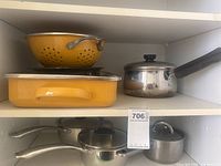 Overall view: yellow enamel colander, roasting pan, three stainless saucepans with lids on shelving