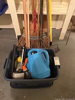 Overview of tote with watering can and long-handle tools behind (rakes, snow shovel)