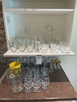 Cabinet shelf showing tumbler glasses, pitcher, dessert dishes, rocks glasses, pepper grinder and goblets below
