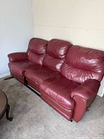 Front angled view of maroon leather reclining three-seat sofa showing torn stitching and reclining mechanism rail
