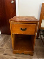 Small wooden side table featuring single drawer with metal bail pull and open lower shelf, visible surface wear