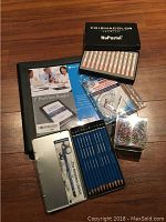 Photo of art supplies including pencils, pastels, binder, protractor, paper clips, and staples on a wooden surface