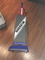 Front view showing upright vacuum with grey Oreck bag and red power cord wrapped around handle