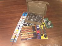 Overview of security sensor kit components and home tools spread on wooden floor, including drain snake, razor blades, utility knives, and LED light puck.