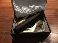 Photo showing gold high heel pumps inside original black shoe box with tissue paper.