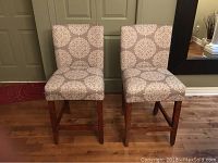 Two bar height chairs side by side showing front and side profile, upholstered in beige fabric with white medallion pattern and wood legs.