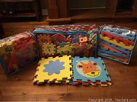 Overall view of three clear storage bags filled with colorful interlocking sponge puzzle mats, plus two example pieces laid flat showing a blue flower and orange giraffe face design.