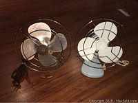 Two vintage electric fans on wood floor, showing their front protective wire cages and metal blades.