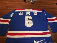 Full view of back of blue hockey jersey with large white number 6, Korean player name, red and white stripes, Nike logo, and visible signatures on fabric.
