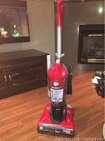Full view of Dirt Devil Featherlite vacuum cleaner upright on a wooden floor with TV and fireplace in background.