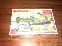 Top view of the closed model kit box showing artwork of Grumman Hellcat aircraft flying over a carrier deck with explosions in background.