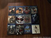 Front view of 12 Blu-ray movies with full covers visible on a wooden floor; movies include Batman Begins, The Dark Knight Rises, Spider-Man, Riddick, Man of Steel, Saw III, Avatar, Pacific Rim, Lords of Salem, Labyrinth, and Looper.