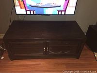 Front view of dark brown wood cabinet with two doors and glass panels showing shelves, placed in front of TV on wood floor.